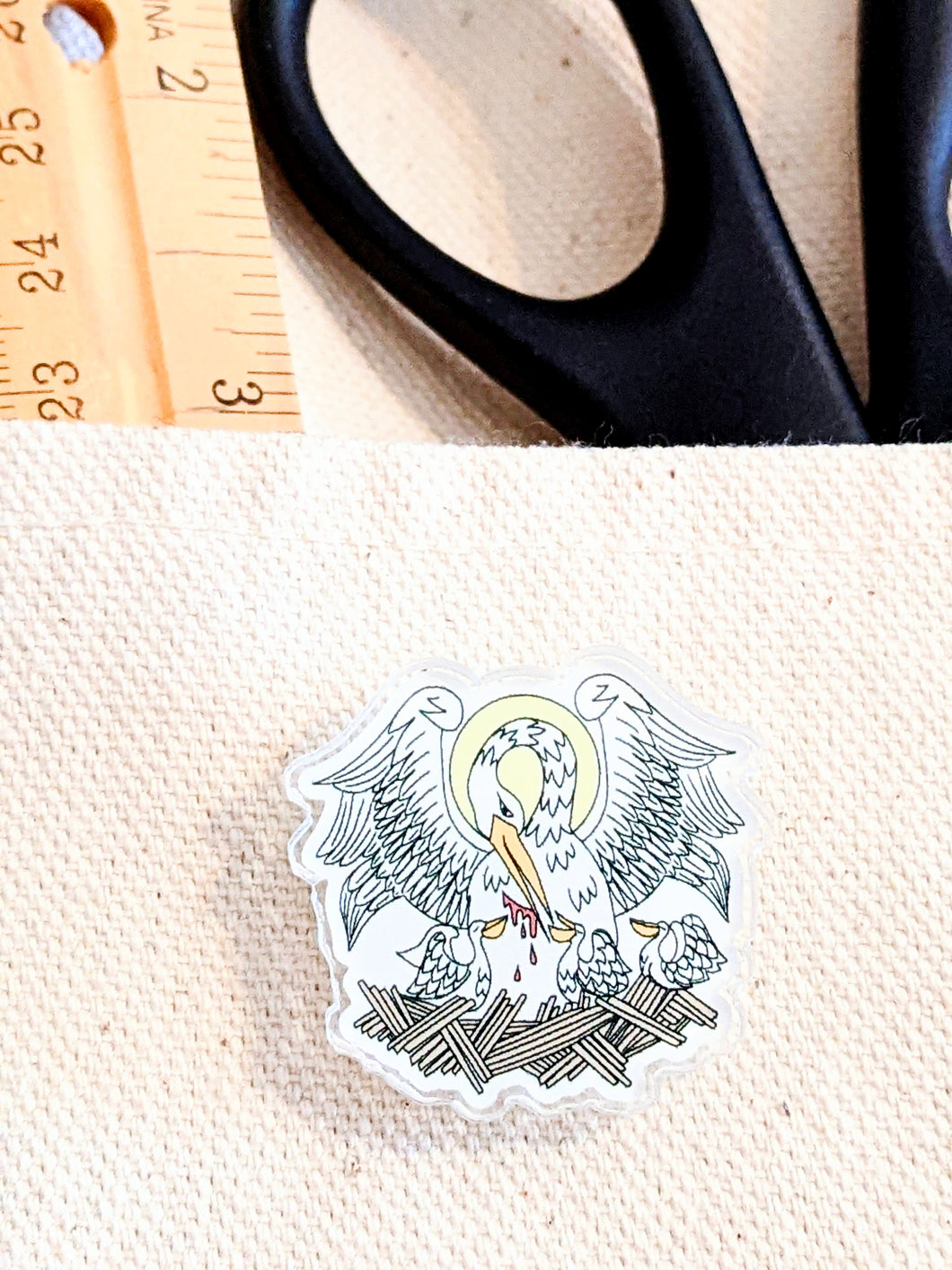 A pelican in her peity acrylic pin is attached to a canvas bag with a ruler and black handled scissors in the backgroun