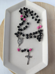 A black rosary with hot pink skulls lays on a white tray. Features a St Joseph Centerpiece and St Benedict Crucifix. 