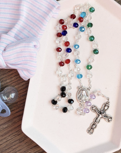 A rosary featuring red, clear. Vlue, aqua, and black beads lays against a white background with a babys hospital cap and pacifier lying next to it. 