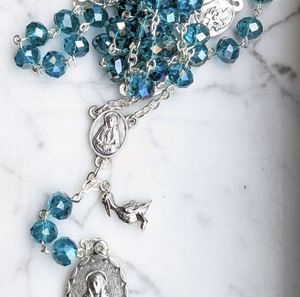 A teal 7 sorrows chaplet with a small pelican charm lays on a marble background 