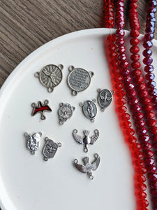 An assortment of Confirmation/ holy spirit themed centerpieces lay on a white tray with 4 strands of different red glass beads next to it. There is a wooden background behind the tray 