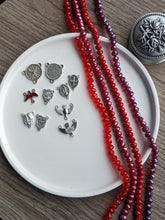 Load image into Gallery viewer, An assortment of rosary centerpieces lay next to 4 strands of red glass beads on a white tray. The tray has a small medal rose rosary box next to it on a wooden background. 