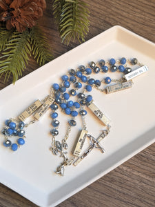 A blie and silver mysteries rosary lays in a white tray with a wooden and fir background