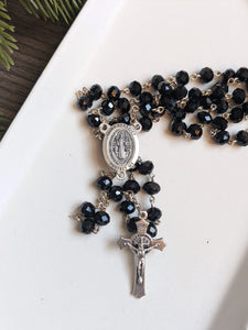 a close up view of the Black St benedict rosary centerpiece and crucifix on a white tray with a wood and fir tree background