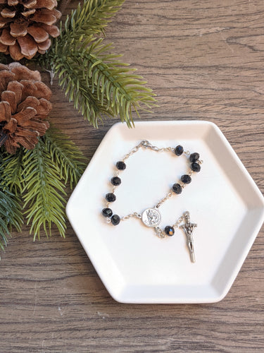 A black glass bead xar rosary with a St Christopher centerpiece and jpII style crucifix lays on a white tray with a wood background and decorative plant