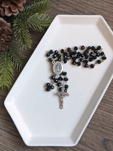 a black glass bead rosary sits on a white tray with a pine branch and wooden background